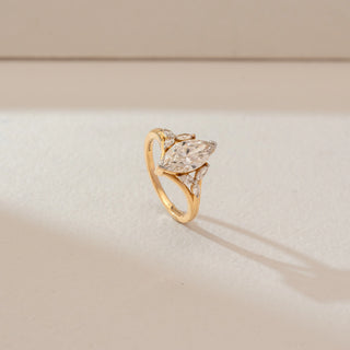 Angled view of marquise diamond ring in yellow gold, featuring a central lab-grown diamond with marquise side stones along a sleek band. Captured against a soft beige textured background.