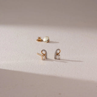Top angled view of diamond stud earrings in 9KT yellow gold, featuring curved pavé loops with baguette-cut diamond accents, set on a beige surface highlighting modern design and subtle sparkle.
