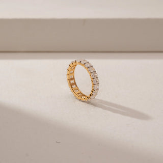 Angled view of a oval diamond eternity ring in 14KT Yellow Gold, featuring a continuous row of oval diamonds against a beige backdrop.