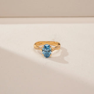 Close up view of a 14KT Yellow Gold diamond ring, featuring an oval blue centre stone and diamond-set shoulders.