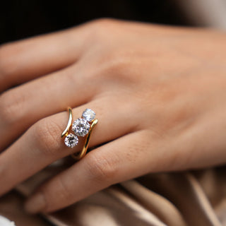 A zoomed in version of an elegant three-stone diamond ring features three sparkling round-cut lab-grown diamonds, set in a unique curved gold band. The diamonds are arranged in a staggered pattern, creating an eye-catching design. The ring is crafted from high-quality gold, ensuring durability and a timeless luster. Close-up view of a model's hand, the Diamond female ring of size 14 boasts 1.9 carats of stunning lab-grown diamonds, set in 18 KT yellow gold weighing a total of 4.52 grams