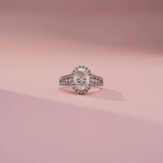Front view of an oval halo engagement ring in White Gold, featuring an oval-cut lab-grown diamond encircled by a sparkling halo and set on a split pavé diamond band, displayed against a soft pink backdrop.