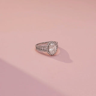 Side view of an oval halo engagement ring in White Gold, featuring an oval-cut lab-grown diamond encircled by a sparkling halo and set on a split pavé diamond band, displayed against a soft pink backdrop.