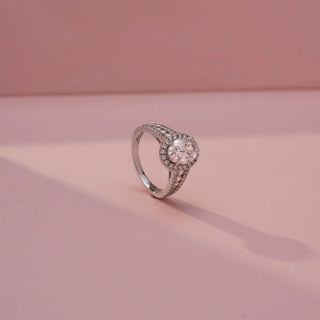 Angled view of an oval halo engagement ring in White Gold, featuring an oval-cut lab-grown diamond encircled by a sparkling halo and set on a split pavé diamond band, displayed against a soft pink backdrop.
