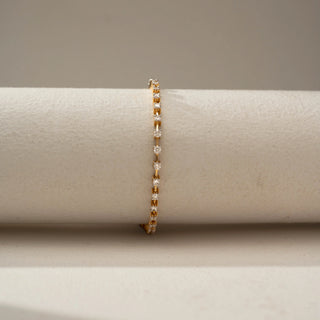 Front view of a delicate gold tennis bracelet featuring a continuous row of round sparkling stones in individual settings, displayed on a neutral cylindrical stand.