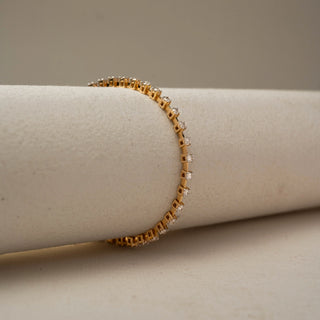 Side view of a gold tennis bracelet showcasing the profile of individual gemstone settings, wrapped around a neutral cylindrical display stand with subtle sparkle and elegant detailing.
