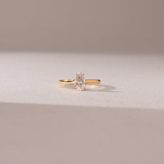 Front view of a diamond ring in 14KT yellow gold, featuring a pear-shaped center diamond in a prong setting on a sleek band, displayed on a beige surface highlighting elegance and brilliance.
