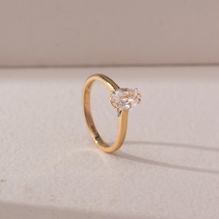 Angled view of a diamond ring in 14KT yellow gold, featuring a pear-shaped center diamond in a prong setting on a sleek band, displayed on a beige surface highlighting brilliance and elegance.
