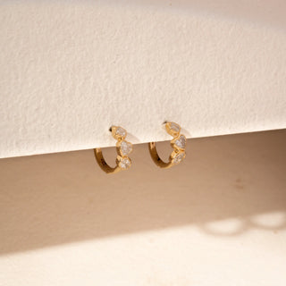 Angled view of diamond hoop earrings in 14KT Yellow Gold, featuring heart, oval and geometric bezel-set diamonds against a beige backdrop.