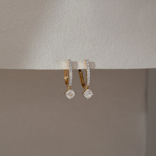 Front angled view of diamond hoop drop earrings in yellow gold, featuring round lab-grown diamonds along the hoop with a square-cut diamond drop. Set against a dual-tone neutral backdrop, they reflect refined sparkle.