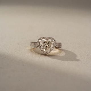 Front view of a heart halo engagement ring in White Gold, featuring a heart-shaped lab-grown diamond encircled by a sparkling halo and set on a triple-row pavé diamond band, displayed against a beige backdrop.