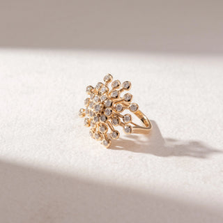 Angled view of floral diamond cluster ring in yellow gold with multiple round diamonds in a radiating design, set on a soft neutral background, highlighting intricate detail and brilliant sparkle.
