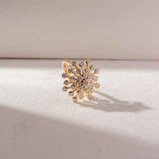 Side view of floral diamond cluster ring in yellow gold with multiple round diamonds in a radiating burst design, set on a soft neutral background, highlighting bold form and radiant sparkle.
