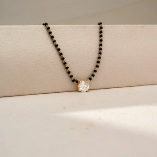 Angled view of a solitaire diamond mangalsutra necklace in 14 KT Dual Gold, featuring a round lab-grown diamond in a classic four-prong setting, suspended from a black bead chain and displayed on a beige backdrop.