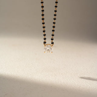 Close Up view of a solitaire diamond mangalsutra necklace in 14 KT Dual Gold, featuring a round lab-grown diamond in a classic four-prong setting, suspended from a black bead chain and displayed on a beige backdrop.