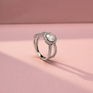 Angled view of a round halo engagement ring in White Gold, featuring a round lab-grown diamond encircled by a sparkling halo and set on a split pavé diamond band, displayed against a soft pink backdrop.