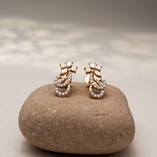 Front view of floral diamond stud earrings in 14 KT Yellow Gold, featuring pavé-set infinity loops with marquise and pear-shaped diamond accents, displayed on a textured stone surface.
