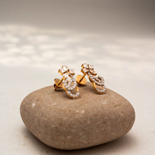 Side view of floral diamond stud earrings in 14 KT Yellow Gold, featuring pavé-set infinity loops with marquise and pear-shaped diamond accents, displayed on a textured stone surface.
