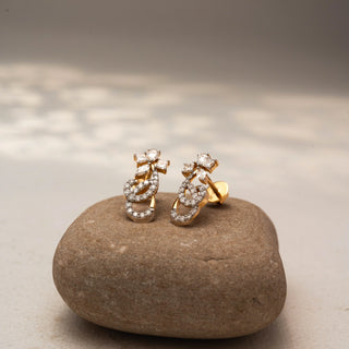Angled view of floral diamond stud earrings in 14 KT Yellow Gold, featuring pavé-set infinity loops with marquise and pear-shaped diamond accents, displayed on a textured stone surface.

