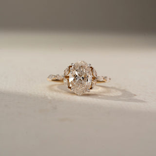 Front view of an oval engagement ring in 14 KT Yellow Gold, featuring an oval-cut lab-grown diamond flanked by marquise-cut side diamonds on a delicate diamond-accented band, displayed against a beige backdrop.