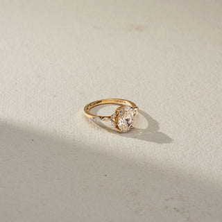 Side view of an oval engagement ring in 14 KT Yellow Gold, featuring an oval-cut lab-grown diamond flanked by marquise-cut side diamonds on a delicate diamond-accented band, displayed against a beige backdrop.