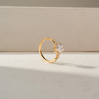 Angled view of an oval engagement ring in 14 KT Yellow Gold, featuring an oval-cut lab-grown diamond flanked by marquise-cut side diamonds in an elevated gallery setting, displayed against a beige backdrop.