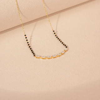 Angled view of a 14KT yellow gold mangalsutra featuring alternating oval, pear and emerald-cut diamonds with black beads, set against a beige background.