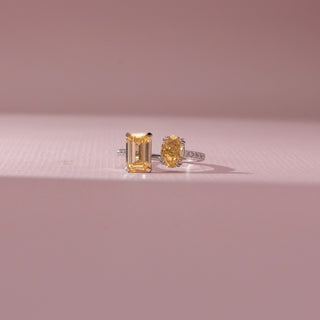 Front view of two yellow diamond rings, one with an emerald-cut stone and the other oval-shaped, set on delicate bands with pavé accents, placed against a soft pink background highlighting warm brilliance.
