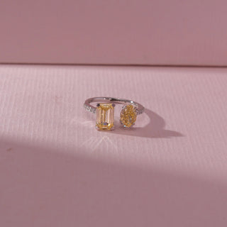 Top view of toi et moi ring with emerald-cut and oval yellow diamonds set on a slim pavé band, placed on a soft pink textured surface, highlighting contrast in shapes and warm radiant sparkle.
