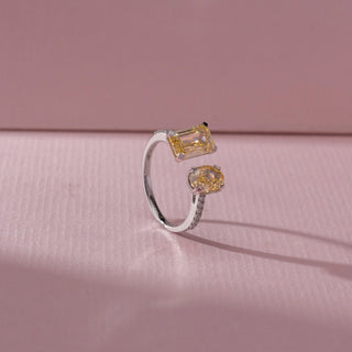 Angled view of toi et moi ring with emerald-cut and oval yellow diamonds on an open pavé band, set against a soft pink textured background, highlighting airy design and warm luminous sparkle.
