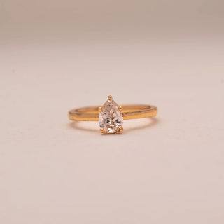 Front view of Reia’s 14 KT Yellow Gold Classic Pear Diamond Promise Ring. The centred pear-shaped solitaire and polished gold band are showcased against a soft pink surface.