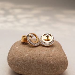 Angled view of circular diamond stud earrings in 14 KT Yellow Gold, featuring pavé-set crescent accents with bezel-set round diamonds, displayed on a textured stone surface.