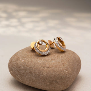 Side view of circular diamond stud earrings in 14 KT Yellow Gold, featuring pavé-set crescent accents with bezel-set round diamonds, displayed on a textured stone surface.