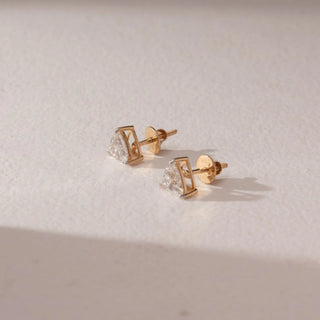 Side view of diamond stud earrings in 14KT yellow gold, featuring trilliant-cut diamonds in prong settings with screw backs, set on a beige surface highlighting structure and brilliance.
