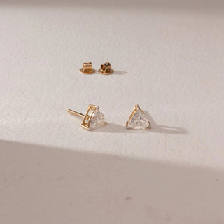 Angled view of diamond stud earrings in 14KT yellow gold, featuring trilliant-cut diamonds in prong settings with screw backs, placed on a beige surface highlighting fine detail and sparkle.
