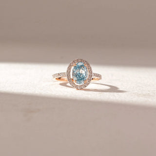 Front view of oval blue gemstone ring in yellow gold, encircled by a pavé diamond halo with a slim pavé band, set on a soft neutral surface, highlighting vivid color and radiant sparkle.
