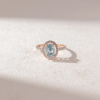 Top view of oval blue gemstone ring in yellow gold, framed by a pavé diamond halo with a slim pavé band, set on a soft neutral surface, highlighting cool tones and refined sparkle.
