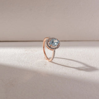 Angled view of oval blue gemstone ring in yellow gold, surrounded by a pavé diamond halo with a slim pavé band, set on a soft neutral background, highlighting airy design and luminous sparkle.
