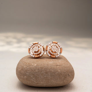Front view of circular diamond stud earrings in 14 KT Rose Gold, featuring concentric pavé-set rings with clustered center diamonds, displayed on a textured stone surface.