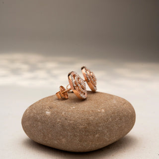 Side view of circular diamond stud earrings in 14 KT Rose Gold, featuring concentric pavé-set rings with clustered center diamonds, displayed on a textured stone surface.
