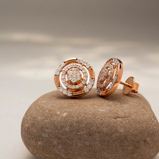 Angled view of circular diamond stud earrings in 14 KT Rose Gold, featuring concentric pavé-set rings with clustered center diamonds, displayed on a textured stone surface.