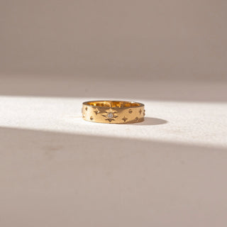 Front view of Stellar Glow Wide in 14KT Yellow Gold, featuring engraved star patterns with a central diamond accent. Set on a soft beige surface, the design reflects subtle texture and modern charm.
