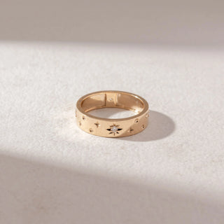 Top view of Star Motif Band Ring in 14KT Yellow Gold, featuring engraved star patterns with a central diamond accent. Set on a soft beige surface, the design highlights subtle texture and shine.
