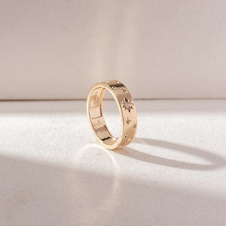 Angled view of Star Motif Band Ring in 14KT Yellow Gold, featuring engraved star details with a central diamond accent. Set on a soft beige surface, the design showcases texture and subtle brilliance.
