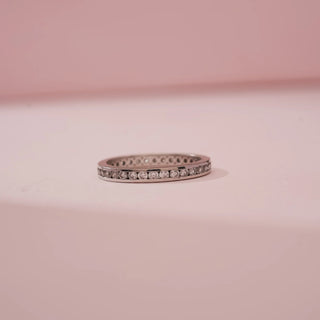 Front view of Reia’s 14 KT White Gold Everyday Luxe Diamond Band Ring. It features a continuous row of shimmering round brilliant diamonds in a channel setting against a soft pink backdrop.