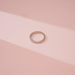 Top view of Reia’s 14 KT White Gold Everyday Luxe Diamond Band Ring. It showcases the slim, polished band and the continuous row of pavé-set round diamonds against a soft pink backdrop.