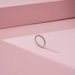 Angled view of Reia’s 14 KT White Gold Everyday Luxe Diamond Band Ring. It features a continuous row of sparkling round brilliant pavé-set diamonds on a slim band against a soft pink backdrop.