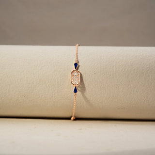 Front view of a diamond bracelet in 14 KT Rose Gold, featuring an oval diamond cluster centerpiece accented with deep blue geometric gemstones on a delicate chain, displayed against a soft beige backdrop.

