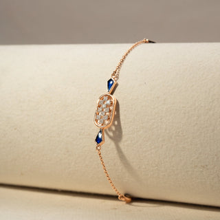 Close Up view of a diamond bracelet in 14 KT Rose Gold, featuring an oval diamond cluster centerpiece accented with deep blue geometric gemstones on a delicate chain, displayed against a soft beige backdrop.
