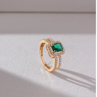 Side angled view of emerald centre halo ring in yellow gold, featuring a rectangular green gemstone framed by round lab-grown diamonds with a split diamond-studded band. Set against a soft neutral backdrop, it highlights structure and sparkle.